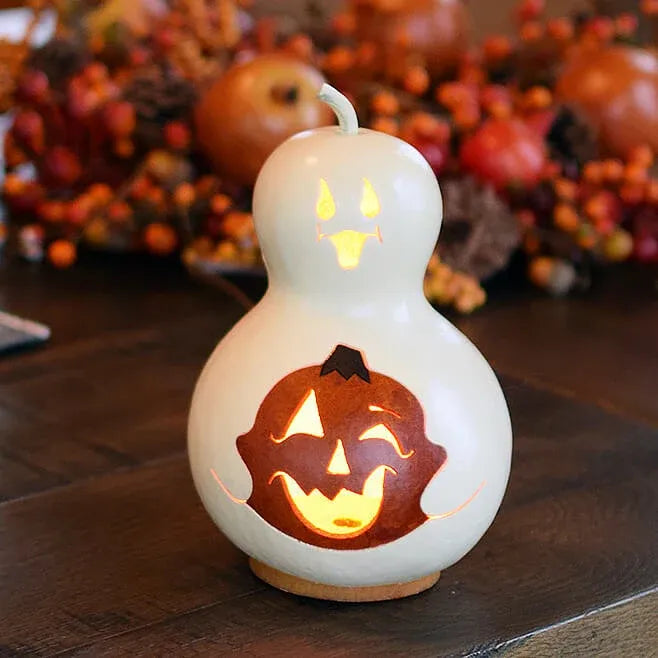 A white gourd-shaped candle with a carved jack-o'-lantern face, glowing with warm yellow light, set on a dark wooden table with autumn decorations blurred in the background.