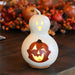A white gourd-shaped candle with a carved jack-o'-lantern face, glowing with warm yellow light, set on a dark wooden table with autumn decorations blurred in the background.