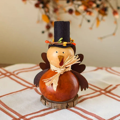 A decorative Thanksgiving turkey ornament made from a gourd, with a black felt hat, painted brown wings, a tan face, and a straw-like scarf, displayed on a plaid tablecloth.