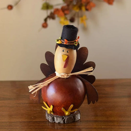 A decorative Thanksgiving turkey figurine made from a gourd and wood slice, wearing a black hat with orange and yellow accents, standing on a small bark base on a wooden table.