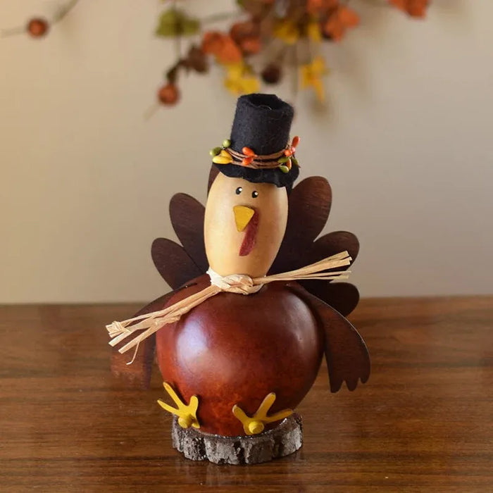 A decorative Thanksgiving turkey figurine made from a gourd and wood slice, wearing a black hat with orange and yellow accents, standing on a small bark base on a wooden table.