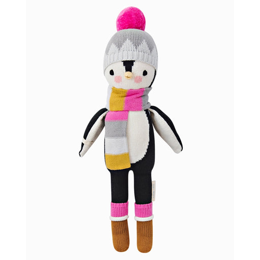 Cuddle + Kind Aspen the Penguin doll standing upright, wearing a gray knit hat, colorful striped scarf, and brown boots.