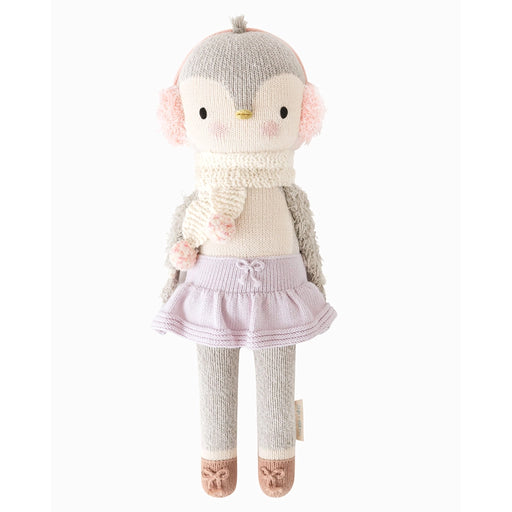 Front view of Cuddle + Kind Skylar the Penguin doll wearing soft pink earmuffs, a cream knit scarf with pom-poms, lavender skirt, gray leggings, and brown knit shoes.