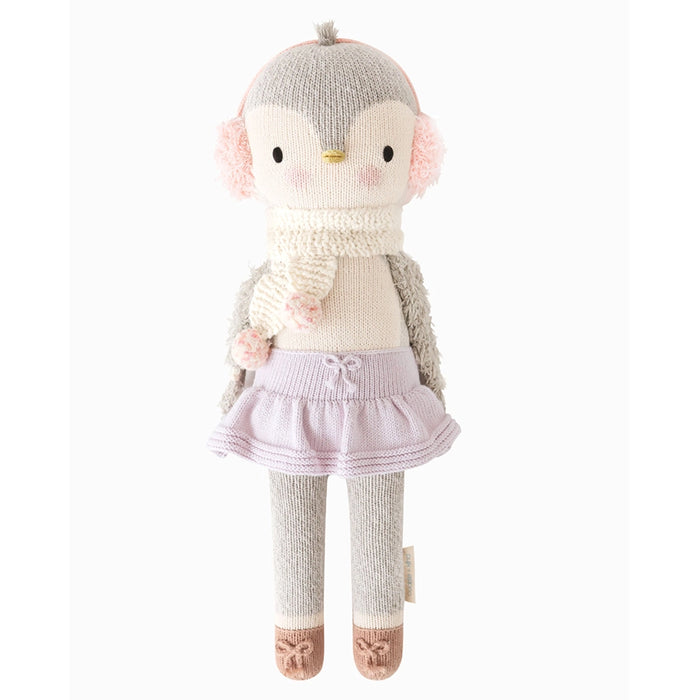 Front view of Cuddle + Kind Skylar the Penguin doll wearing soft pink earmuffs, a cream knit scarf with pom-poms, lavender skirt, gray leggings, and brown knit shoes.