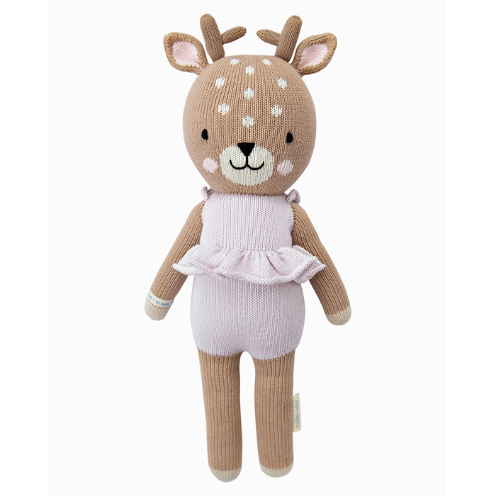 A knitted deer doll wearing a soft lavender romper with a ruffled waist, smiling with white embroidered spots on its head.