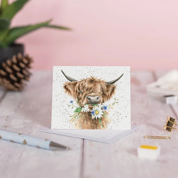 A highland cow illustrated on a greeting card, displaying its iconic shaggy fur and friendly demeanor in a pastoral setting.
