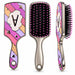 Three views of a decorative paddle hairbrush on a white background: front, side, and back. The brush has a metallic silver handle and head frame, black bristles with pink tips, and a colorful geometric face in pink, purple, orange, and white with a large letter A.