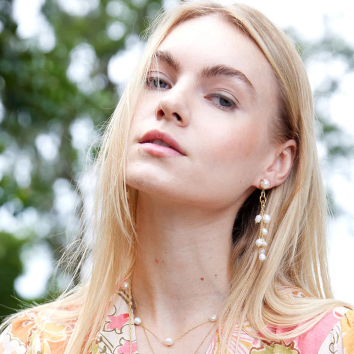 A young woman with long, straight blonde hair poses confidently, wearing floral attire and earrings with pearls.