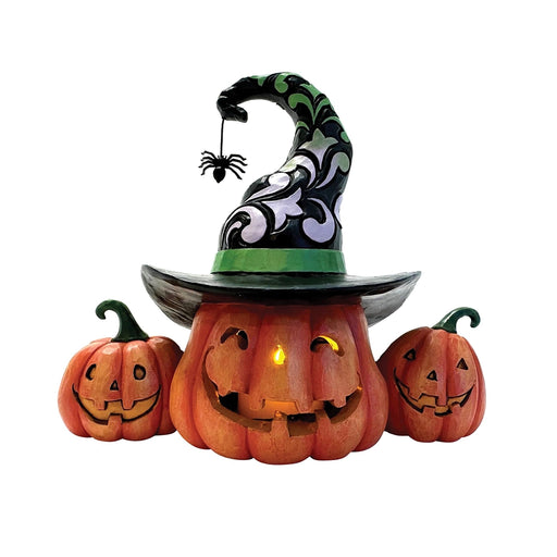 A carved pumpkin wearing a black witch hat with green trim and dangling spider, flanked by two smaller pumpkins.