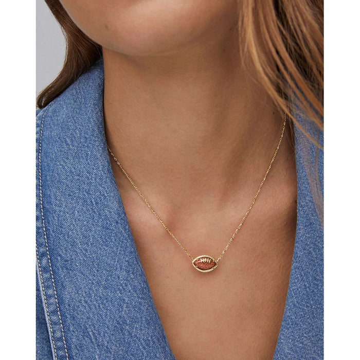 Model wearing Kendra Scott gold football short pendant necklace with orange goldstone charm