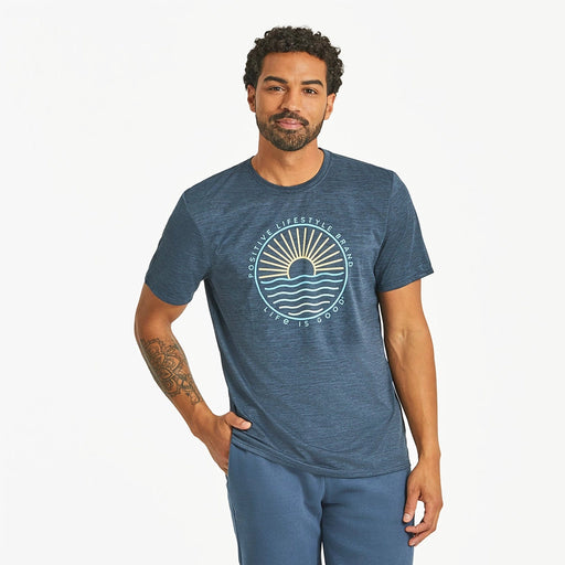 Front view of Life Is Good Men's Sun & Waves Crusher Tee on model – navy blue graphic t-shirt with sunrise and wave design, paired with blue shorts.