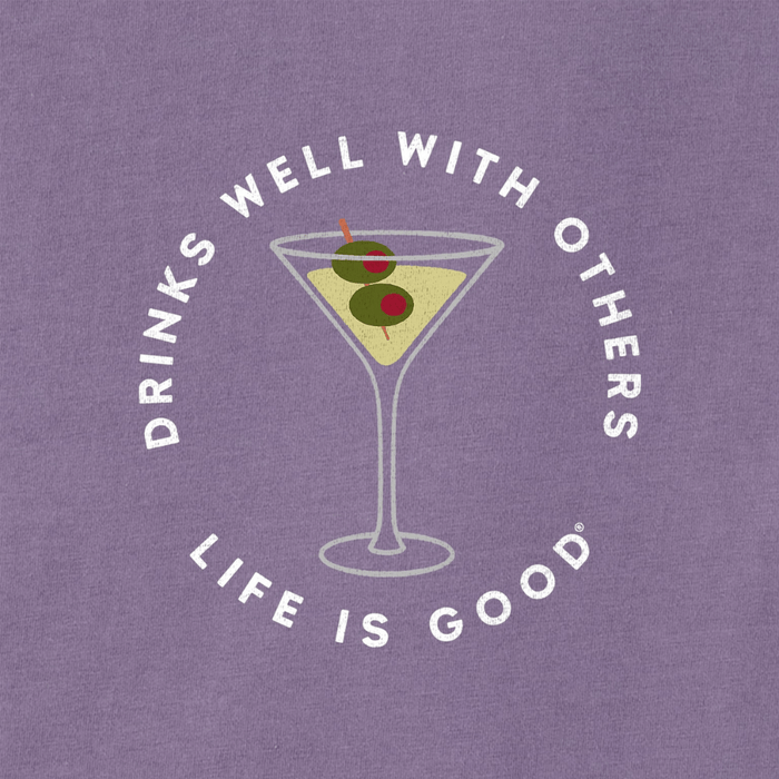 Graphic of a martini glass with green olives and the phrase "Drinks Well With Others" in white text, centered on a purple women's t-shirt with "Life Is Good" branding underneath.