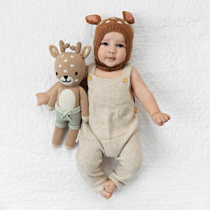 Baby wearing a knitted deer hat and overalls, holding a Little Cuddle + Kind Elliott the Fawn