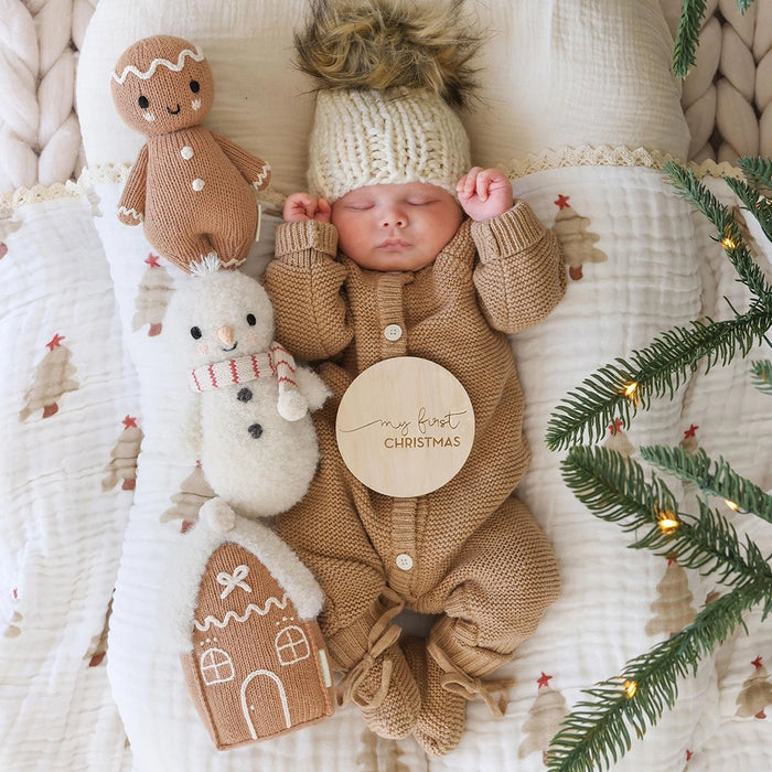 Baby dressed in cozy knit outfit lying with Cuddle + Kind baby snowman, gingerbread doll, and gingerbread house plush, holding a “My First Christmas” sign.