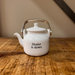 Mud Pie “Shake It Down” white ceramic shaker shaped like a teapot with a metal handle, sitting on a wooden surface.