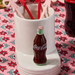 Nora Fleming Coke® Bottle Mini in ceramic inserted in serving base with bow drink stirrers and Coca-Cola décor