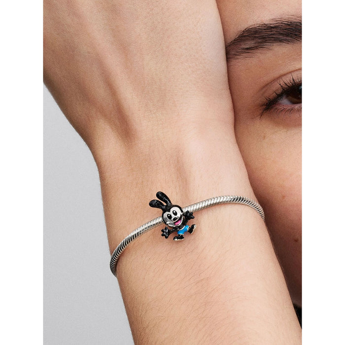 A close-up of a person’s wrist wearing a silver bracelet with the Pandora Disney Oswald the Lucky Rabbit charm. The charm is detailed in black enamel with blue shorts, featuring Oswald’s signature long ears and cheerful pose.