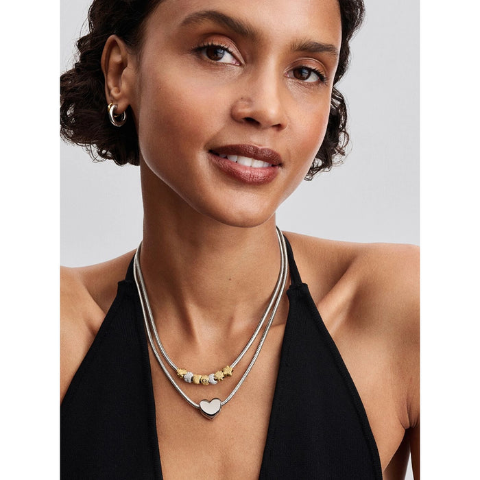A woman wearing layered snake chain necklaces in silver tones, one with a smooth silver heart pendant and another with multiple charms in silver and gold.