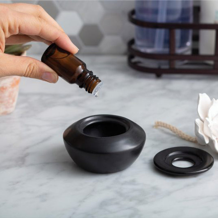 Hand pouring essential oil into a black ceramic diffuser on a marble surface.