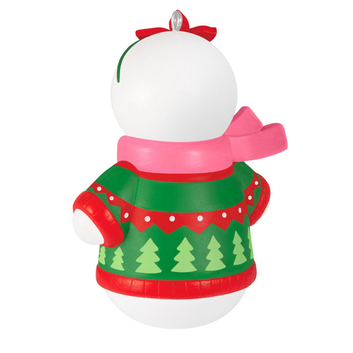 Decorative Christmas ornament with a white ball, red bow, pink scarf, and green sweater with tree pattern on a white background.