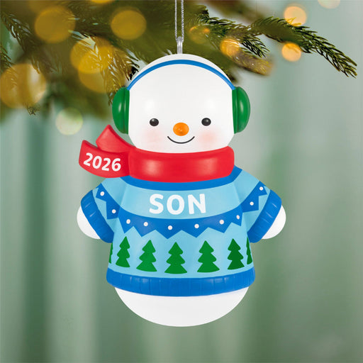 Snowman ornament with a blue sweater, red scarf, and green earmuffs hanging on a Christmas tree.
