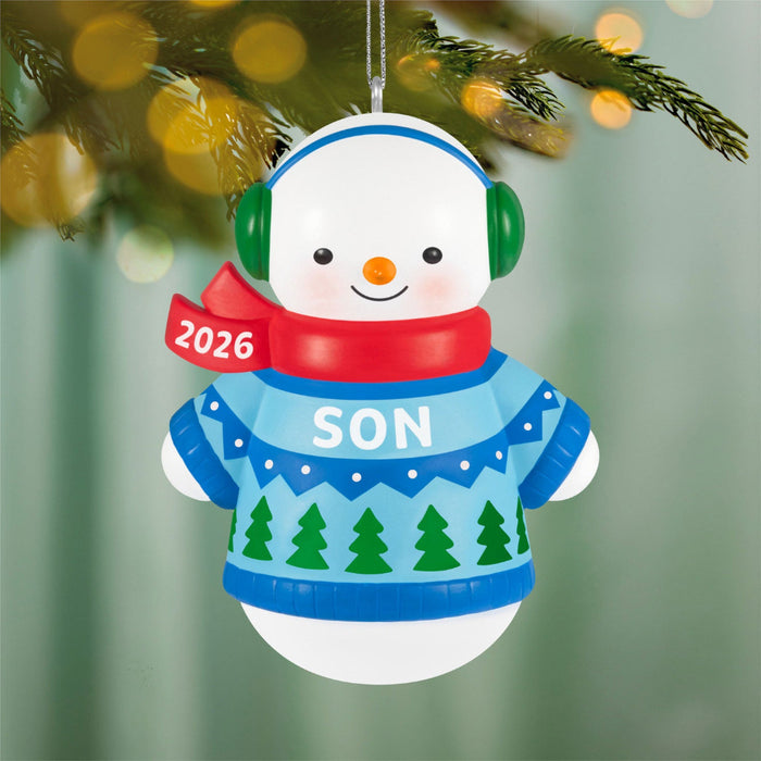 Snowman ornament with a blue sweater, red scarf, and green earmuffs hanging on a Christmas tree.