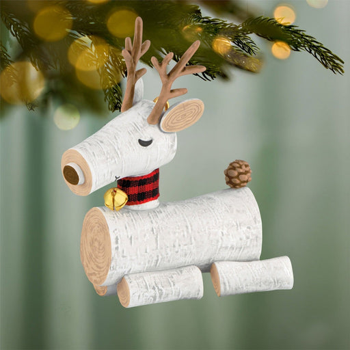 Close-up of rustic birch log reindeer Christmas ornament hanging on green fir branch with gold bell detail