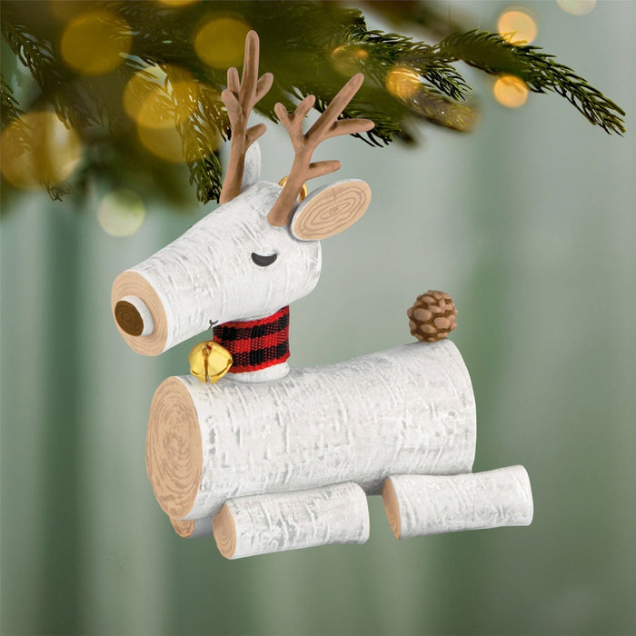 Close-up of rustic birch log reindeer Christmas ornament hanging on green fir branch with gold bell detail