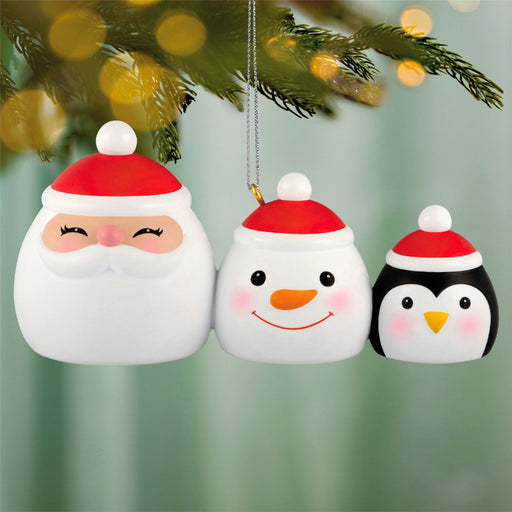 Three Christmas ornaments shaped like a Santa Claus, snowman, and penguin hanging on a tree.