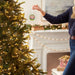 Person decorating a Christmas tree with lights and ornaments in a festive living room.