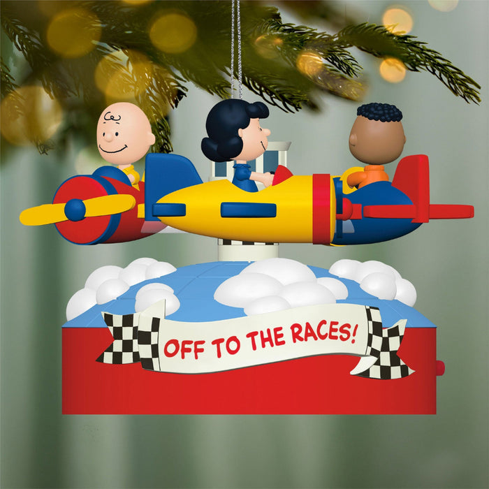 Decorative Christmas ornament with cartoon characters on a race car, 'Off to the Races!' text, and checkered flags against a blurred Christmas tree background.