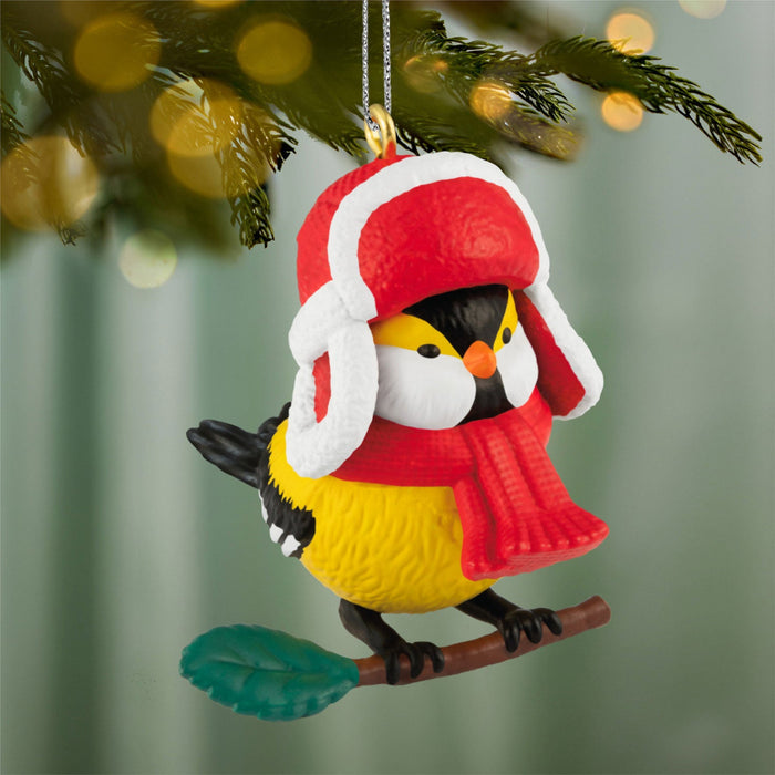 Decorative bird ornament wearing a red hat and scarf on a branch with a blurred Christmas tree background.