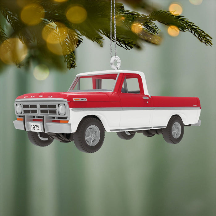 Red and white truck ornament hanging on a Christmas tree branch with blurred lights in the background.
