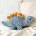 Gray plush stegosaurus toy with golden plates resting on a cream blanket beside a beige cushion
