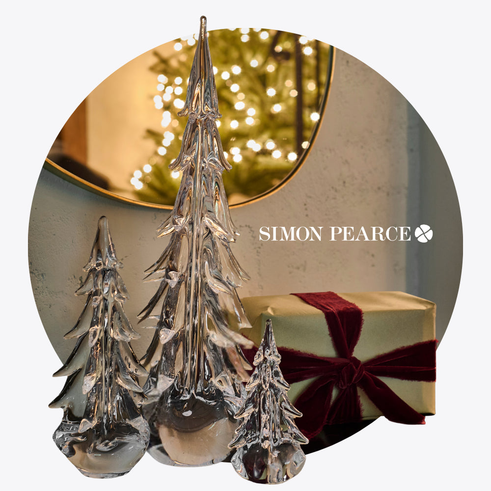 Crystal Christmas trees with a wrapped gift and fairy lights in the background, featuring Simon Pearce logo.
