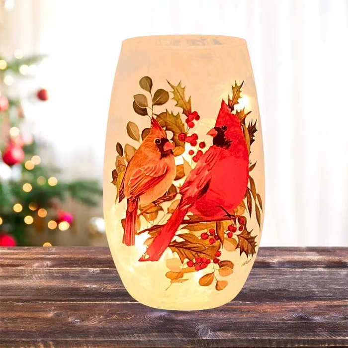 Pre-lit frosted glass vase with two cardinals perched on holly branches glowing warmly on a wood surface.