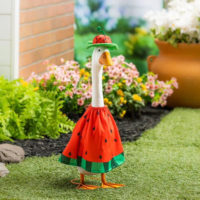 A duck in a watermelon-patterned dress stands proudly in a vibrant garden filled with flowers and greenery.
