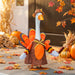 A decorative turkey-shaped garden statue wearing a brown dress and hat, with orange, yellow, and black feather details, standing on a wooden deck surrounded by pumpkins, autumn leaves, and corn in a fall outdoor setting.