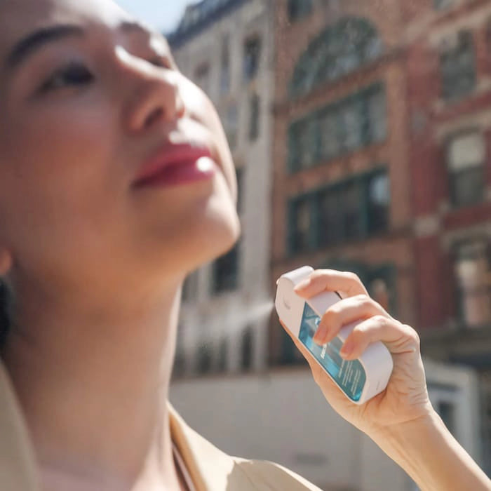 Person applying body mist spray outdoors with buildings in the background