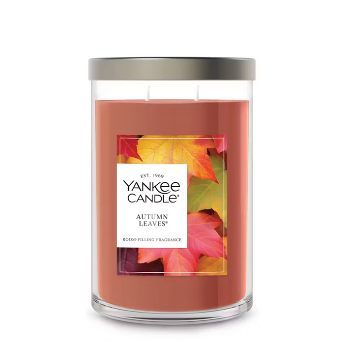 A large Yankee Candle Autumn Leaves tumbler with two lit wicks. The label features colorful autumn leaves in red, orange, and yellow. A real autumn leaf is placed at the base of the candle against a warm background.