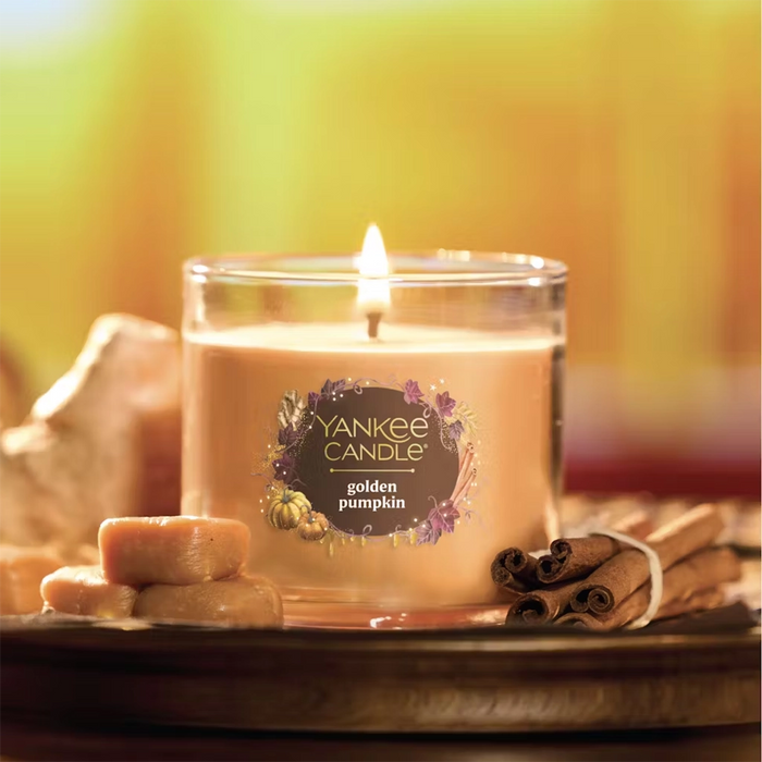 Small Yankee Candle golden pumpkin candle on a wooden surface with cinnamon sticks and apples.