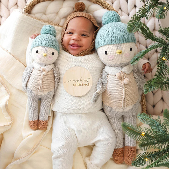 Baby smiling while holding Cuddle + Kind Greyson the Penguin dolls, one Little size and one regular, during first Christmas photo.