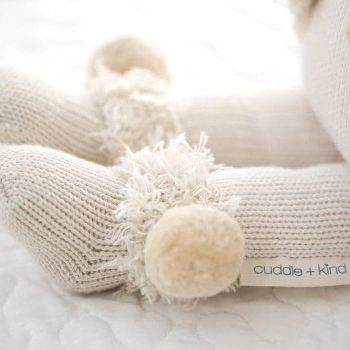 Close-up of the knit details on Stella the Polar Bear doll’s feet with cream pom-poms and the "cuddle + kind" fabric tag visible.
