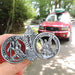 Two silver coins with religious designs held by a hand, with a red vehicle and greenery in the background.