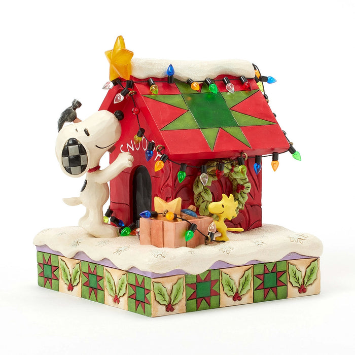 Front view of Snoopy decorating his festive red doghouse with a star and lights, Woodstock nearby with presents.