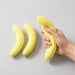 Hand holding a banana next to two bananas on a plate with a wooden tray in the background.