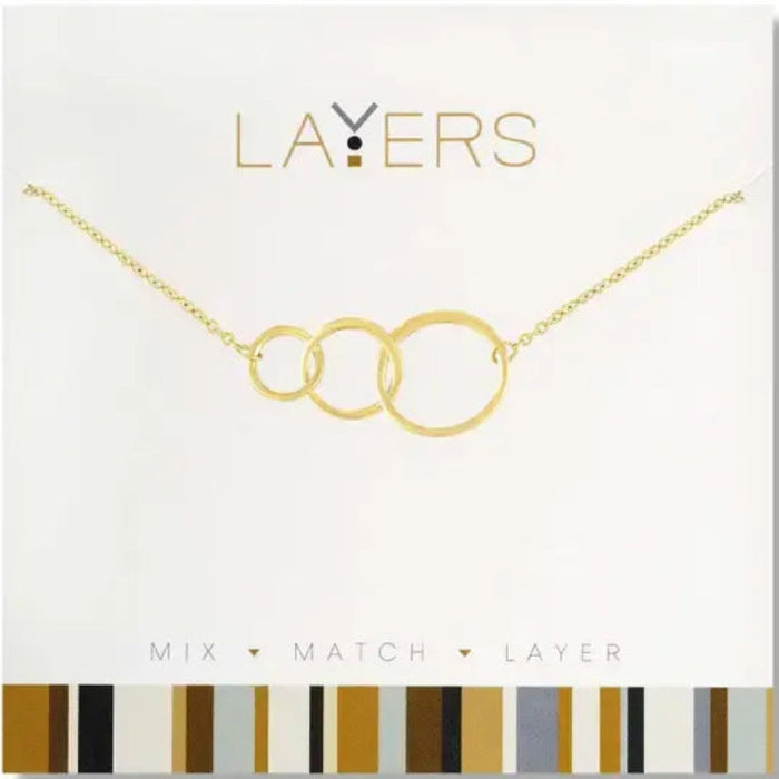 A gold necklace with three interlocking layered rings on a delicate chain, presented on a white card labeled 'LAYERS' with a gold and brown striped pattern at the bottom.