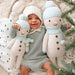 Smiling baby in a knit outfit holding two Spruce the Snowman dolls, one large and one small, while sitting next to a Christmas tree.