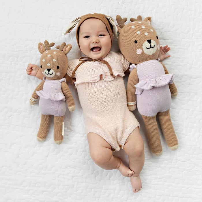 Smiling baby lying on a white blanket, wearing a pale pink romper and holding two lavender romper deer dolls, one small and one large.